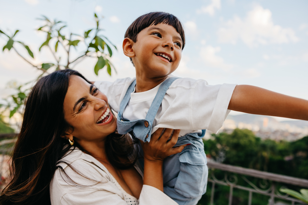 A Latin mother laughing with her young son during a cheerful moment outside, enjoying sunny weather and family time together, featuring natural greenery in the